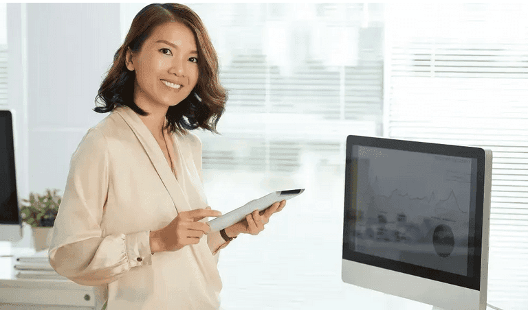 A smiling woman holding a tablet beside a computer screen
