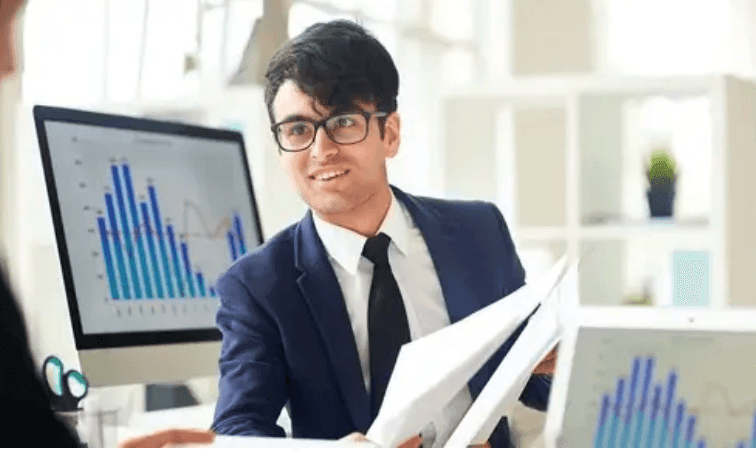 A smiling businessman holding papers in front of screens with bar charts